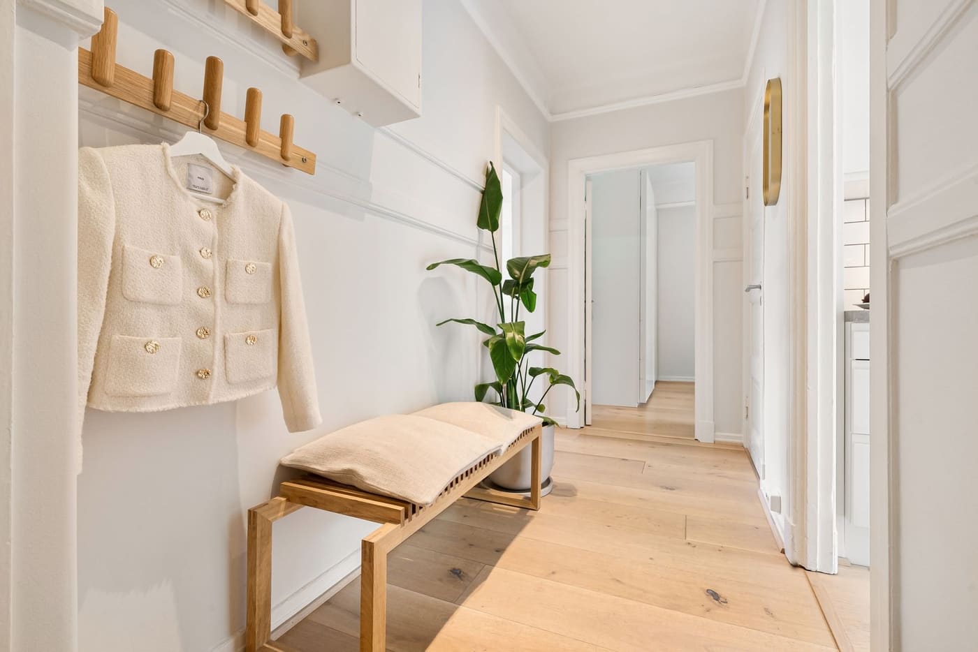 A bright narrow hallway with a wooden bench, a plant and a jacket on a wall rack