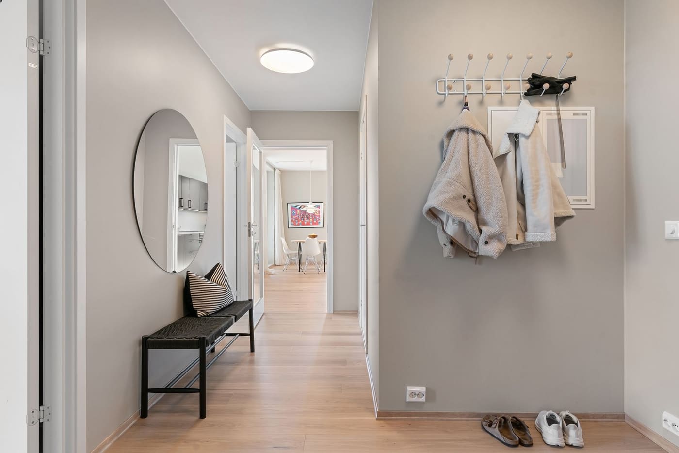 A hallway with coats on wall hooks, a wooden bench with shoes beneath it and a round mirror