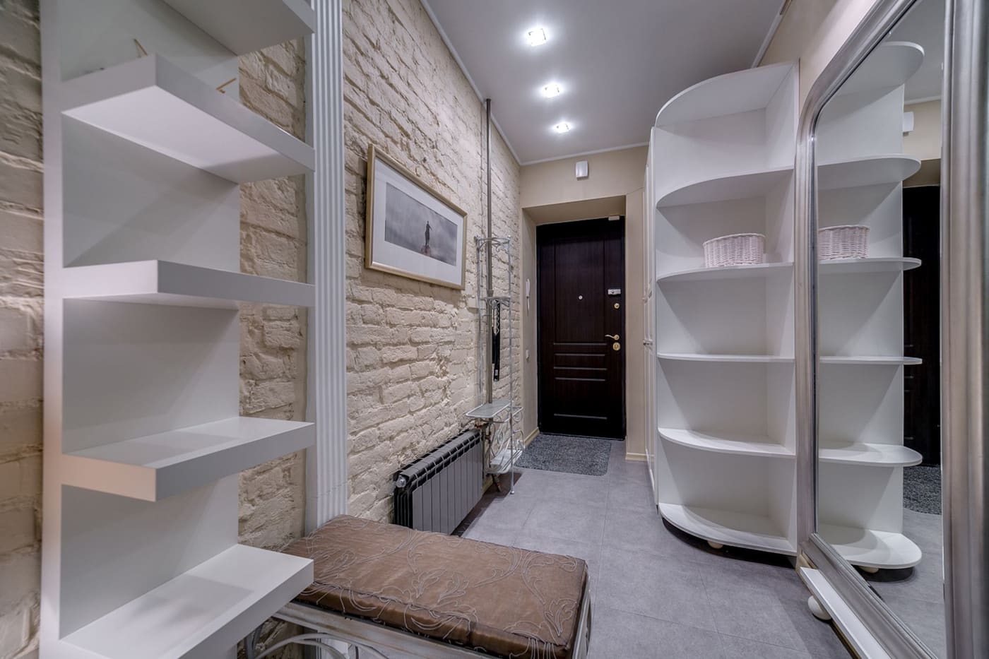 A narrow entry with open white wall shelving, a cushioned bench and a mirrored wall
