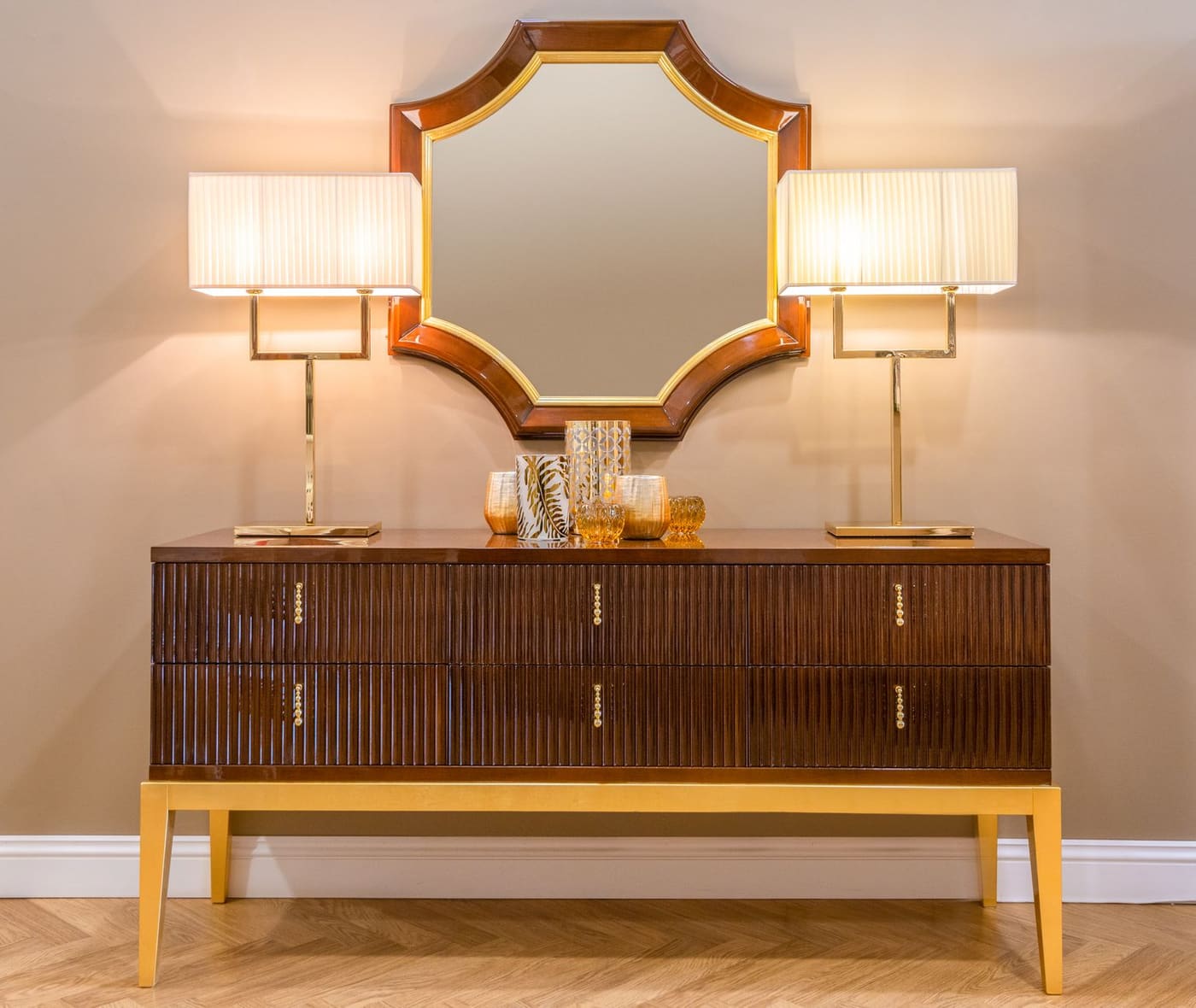 A wooden chest of drawers with two matching table lamps and a gold-framed mirror above